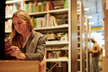 Serious blond woman sitting in front of camera in modern library and reading book against huge stacks of bookshelves