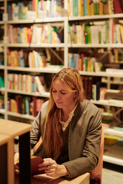 Young female bibliophile in smart casualwear reading book in library while sitting by desk against stack of bookshelves