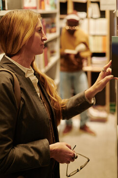 Side view of serious blond woman taking book from bookshelf in library or modern shop against another bibliophile