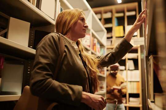Blond female in blazer standing between stacks of shelves in library and taking book while looking for particular one
