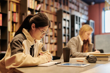 Diligent young woman pointing at page of book while reading it in library at leisure or after classes in college or university