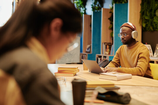 Young serious man in headphones networking by table in front of laptop while sitting in library and preparing for exam