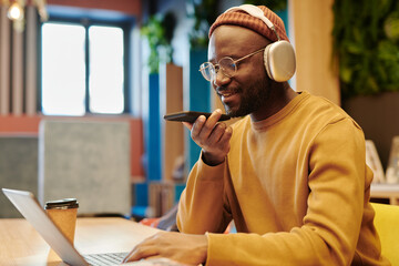 Young smiling male student speaking in mobile phone while recording voice mail and networking in front of laptop