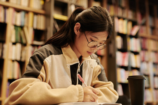 Young Asian female student with pen writing down main points of online book while preparing for seminar in reading hall