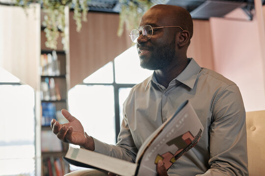 Young African American popular writer with open book talking to public at literature event in modern library