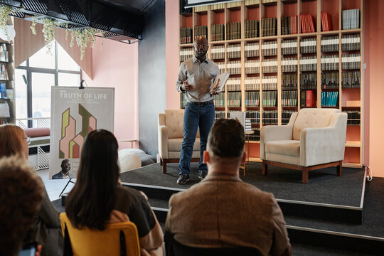 Young writer with his new book standing on stage with armchairs in modern library and communicating with public