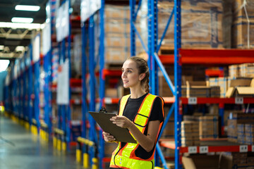 A woman wearing a safety vest and holding a clipboard in a warehouse. She is smiling and looking up...