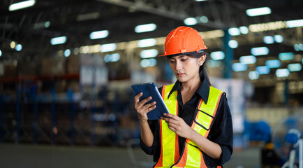 A woman wearing a safety vest and an orange helmet is looking at a tablet. She is likely a worker in a factory or warehouse, using the tablet to check information or instructions - obrazy, fototapety, plakaty