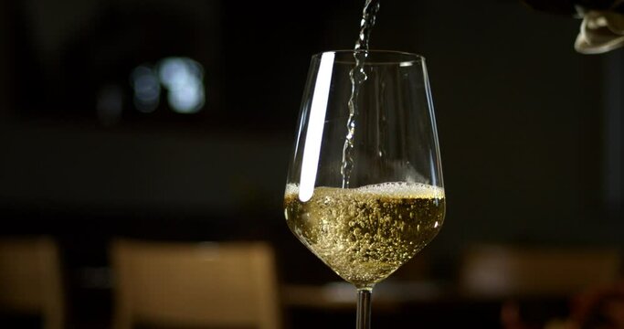 Super slow motion close up of white sparkling wine or champagne is being poured in transparent glass by barman or waiter while serving to customers at dinner at restaurant.