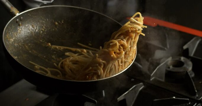 Super slow motion macro of a professional chef is flipping traditional Italian dish of spaghetti with red tomato sauce on pan to be cooked in kitchen of restaurant.