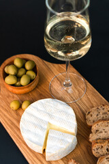 Camembert cheese, crispy baguette, olives and wineglass of white wine on a dark background. Close-up. Selective focus.