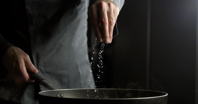 Super slow motion close up of stellar chef is putting heap of granulated rough crystal salt with flying grains while cooking in restaurant kitchen at 1000 fps.