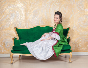 Beautiful smiling woman in green rococo style medieval dress sitting on the sofa