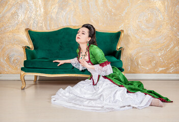 Beautiful sad woman in green rococo style medieval dress sitting on the floor near sofa and raises her hand