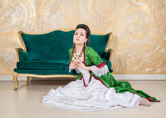 Beautiful sad woman in green rococo style medieval dress sitting on the floor near sofa