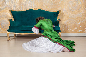 Beautiful sad crying woman in green rococo style medieval dress sitting on the floor near sofa
