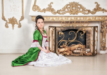Beautiful serious woman in green rococo style medieval dress sitting on the floor near fireplace