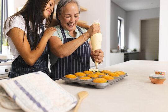 Asian grandmother and biracial teenage granddaughter are decorating cupcakes at home