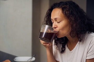 Close-up authentic portrait of a multi ethnic curly haired young pretty woman in pajamas, drinking coffee in the morning. Attractive woman enjoying her espresso coffee during breakfast.