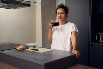 Beautiful positive woman enjoys her morning coffee, smiling looking at camera, standing at the kitchen table