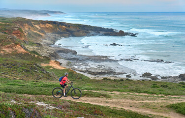 nice senior woman riding her electric mountain bike at the rocky and sandy coastline of the atlantic ocean in Porto Covo, Portugal, Europe - 785612283