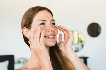 Cheerful young woman taking care of her facial skin, applying moisturizer with SPF effect at home....