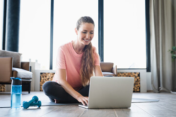 Cheerful young Caucasian woman browsing online on laptop while having training at home. Fit female...