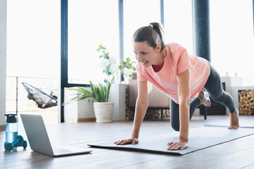 Happy young woman standing in plank position while watching online training on laptop at home....