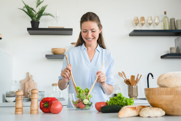 Cheerful young woman mixing vegetables in the bowl at home kitchen. Healthy eating habits concept. Caucasian housewife cooking making salad indoors