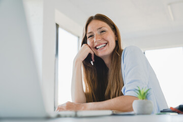 Happy young Caucasian woman working on laptop laughing at office desk during break. Female designer...