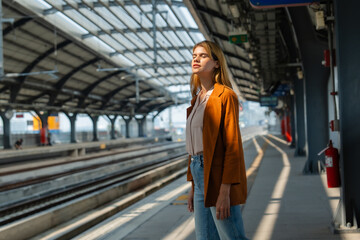 A contemplative young woman stands alone on a sunny train station platform, seemingly waiting for...