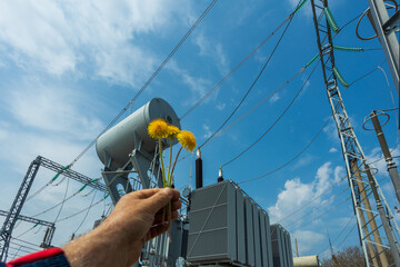 Three-phase high-voltage transformer of high electrical power with high-voltage bushings at a substation against the background of a blue sky and yellow dandelions.