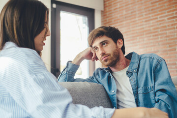 A young 25-year-old couple sits on their cozy sofa in a living room, engaged in a quiet conversation and sharing carefree moments together. Leisure and relaxation concept. Discussion man and woman