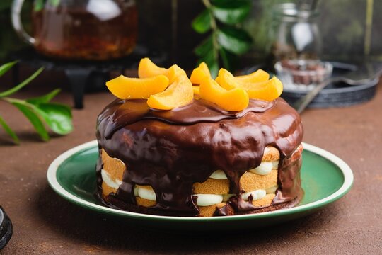 A biscuit cake with a cream of masquearpone, canned peaches covered with chocolate on a green plate on a brown concrete background.