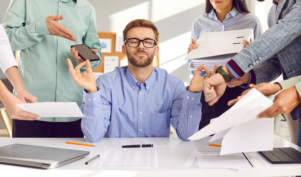 Calm young man in shirt and glasses sitting at working desk with eyes closed, mediating, relaxing, relieving stress, and ignoring team of business colleagues demanding to do lots of office paperwork