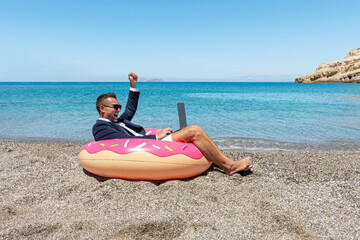 Happy businessman with laptop on inflatable donut on tropical beach. Summer vacation concept.