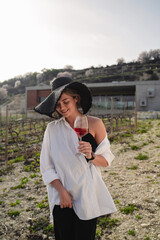Woman in elegant attire and a wide-brimmed hat enjoys a glass of rose wine at a vineyard