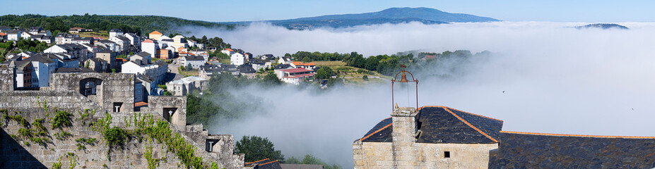 Vista panorámica del pueblo bonito de España Castrocaldelas en Orense entre la niebla blanca con casas, árboles verdes ,montañas y un campanario en primer plano, verano de 2021, España.
