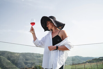 Fototapeta premium Woman in elegant attire and a wide-brimmed hat enjoys a glass of rose wine while overlooking a sprawling vineyard