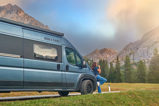 Attractive middle-aged woman leans against motorhome, sipping coffee with Dolomites peaks behind. 