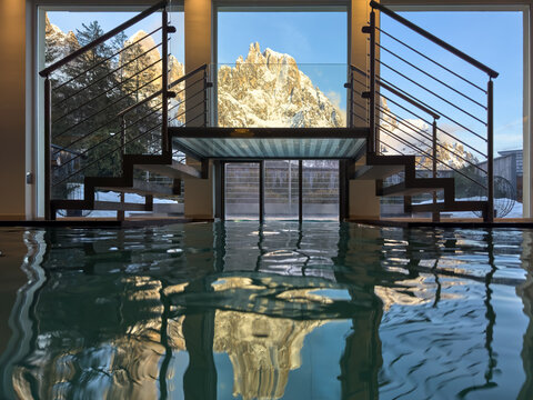 Indoor spa pool with Dolomite mountain panorama framed by glass and stairs &mdash; alpine wellness resort scene. 