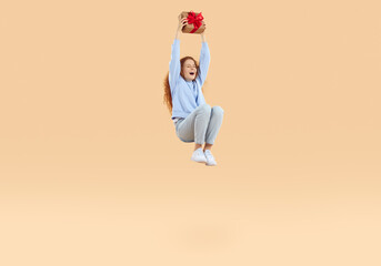Fototapeta na wymiar Happy laughing redhead curly cute girl in blue sweatshirt and jeans is jumping in air with christmas gift box wrapped craft paper with red bow, a present on beige background. Sales, holidays discount.