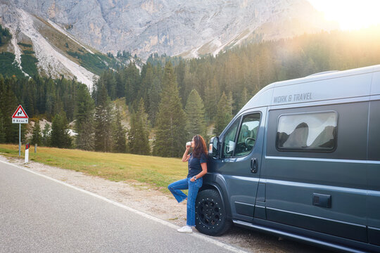An attractive dark-haired woman on an adventure trip in the Dolomites leans against an off-road camper, sipping coffee and watching the mountain peaks illuminated by the setting sun.