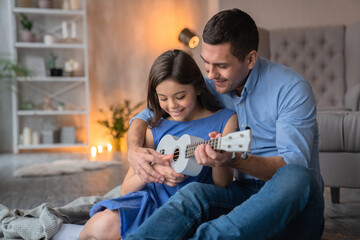 Cute little smiling girl playing on white wooden ukulele guitar with her smiling cheerful daddy dad...