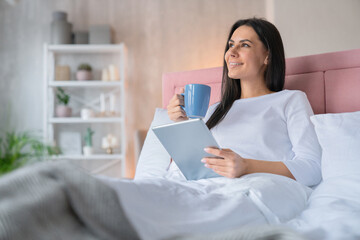 Dreaming moment. Side view shot of young beautiful woman with brown hair in light white pajamas in home bed with cup of morning coffee tea and tablet pad in hands