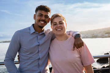 Two friends, male and non-binary, share laughter on a seaside promenade at sunset, with the ocean backdrop highlighting their joyful interaction. Diverse friendship relationship. Waist up.