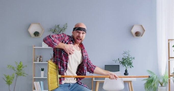 Funny man standing in living room at home playing using mop as a guitar during housework. Young guy having fun while cleaning house. Clean and household concept. 4k video. Slow motion video.