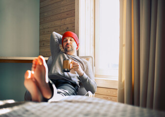 Winter male traveller in red hat holds wooden mug of mulled wine or hot tea with by the window