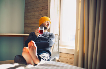 Winter male traveller in yellow hat holds a mug of mulled wine with by the window in wooden cozy...