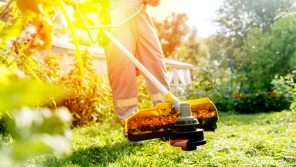 Gordijnen Gardener with trimmer mows lawn in garden © I